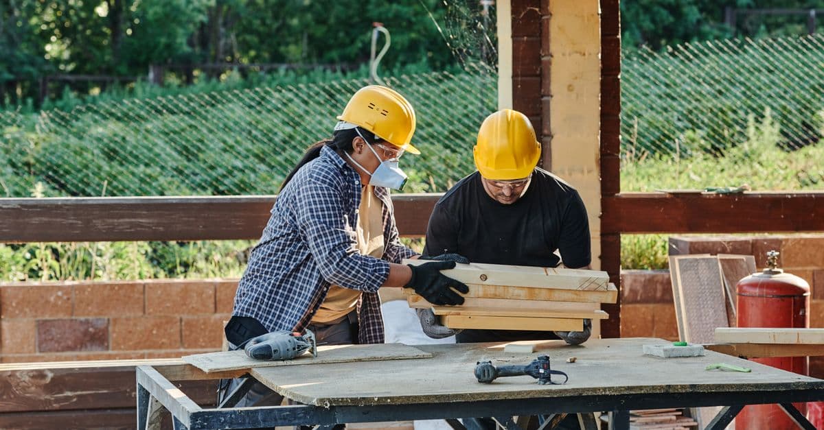 Deux artisans en casque préparant des planches de bois sur un établi de chantier